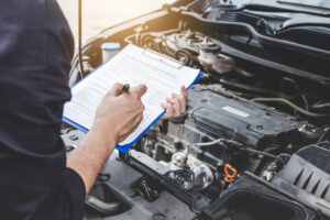 A person holds a pen and clipboard with a maintenance log sheet while looking at a car's engine.